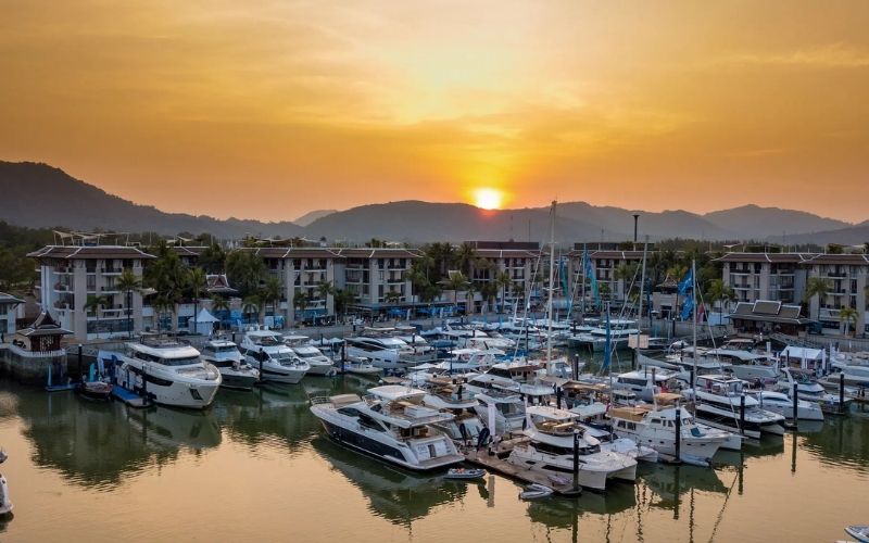 A bright, high-angle aerial photograph of a luxury marina densely packed with white yachts and boats. The marina is surrounded by modern resort buildings, lush green tropical trees, a small inner pond, and distant rolling hills.