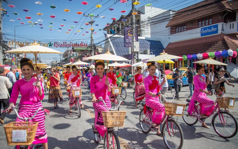 A brightly lit street scene during the Bo Sang Umbrella and Sankhampaeng Craft Festival, showing a parade of young women in matching pink traditional Thai clothing riding bicycles and holding parasols decorated with colorful stripes. The street is lined with spectators and strung with miniature umbrellas overhead.