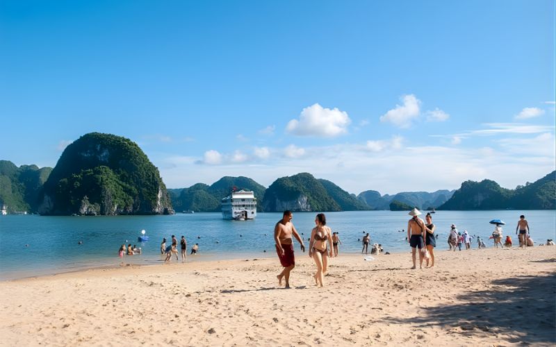 Tourists walking and relaxing on a sandy beach with calm blue water, limestone islands, and a cruise boat in the background under a sunny sky.