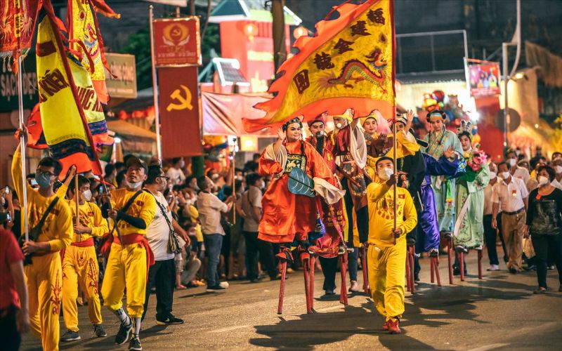 Tet Nguyen Tieu lantern festival street procession in Ho Chi Minh City, Vietnam, featuring performers in traditional colorful costumes walking on stilts, attendants in bright yellow outfits carrying ceremonial flags and banners, and crowds watching along a lively night street decorated with festival lights.
