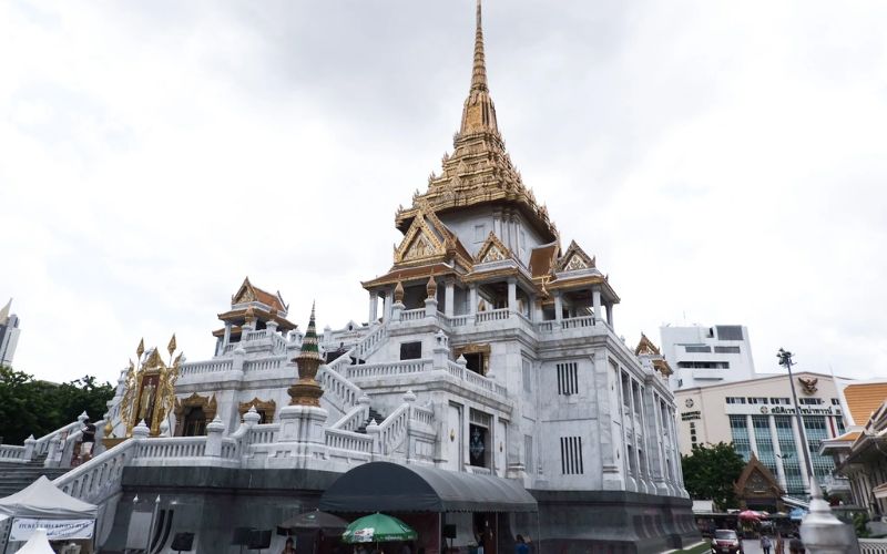 A wide exterior shot of the Phra Maha Mondop at Wat Traimit (The Temple of the Golden Buddha) in Bangkok, featuring a grand four-story white marble building with ornate gold-trimmed gables and a tall, tapering central spire under an overcast sky.