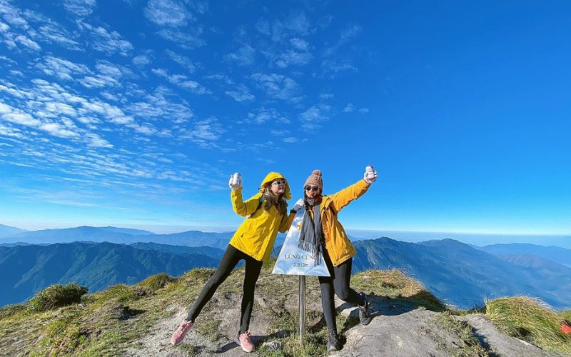 Two hikers in yellow jackets posing joyfully on a mountain peak in Mu Cang Chai, Vietnam, with a clear blue sky and distant mountain ranges in the background