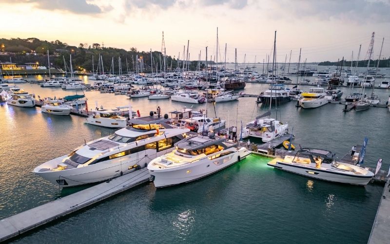 A high-angle view of a luxury marina crowded with large, modern motor yachts and sailboats docked along piers at sunset, promoting the Thailand International Boat Show.