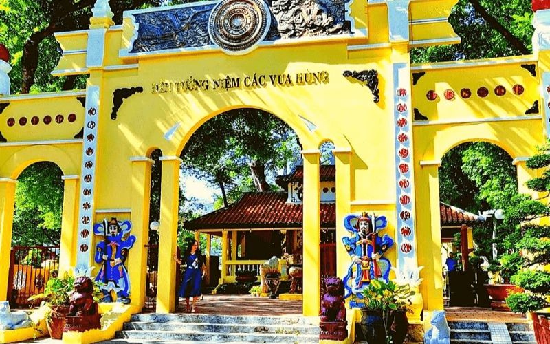 The vibrant yellow ceremonial entrance of the Temple of the Hung Kings in Ho Chi Minh City, featuring traditional Vietnamese triple-gate (Tam Quan) architecture, intricate carvings on the lintel, and two colorful guardian statues flanking the central archway.