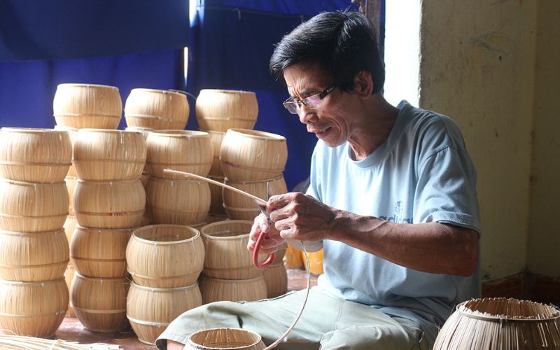 Artisan in a traditional craft village in Vietnam skillfully weaving bamboo into baskets, with stacks of finished products in the background