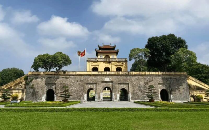 A wide view of the Doan Mon gate at the Imperial Citadel of Thang Long in Hanoi, featuring a massive stone fortress base with arched entrances and a traditional Vietnamese pavilion with a tiered roof on top, set against a backdrop of green lawns and a blue sky.