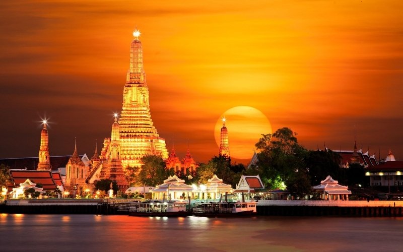 Sunset view of Wat Arun temple in Bangkok, Thailand, with the sky glowing orange and the temple lights reflecting on the Chao Phraya River.