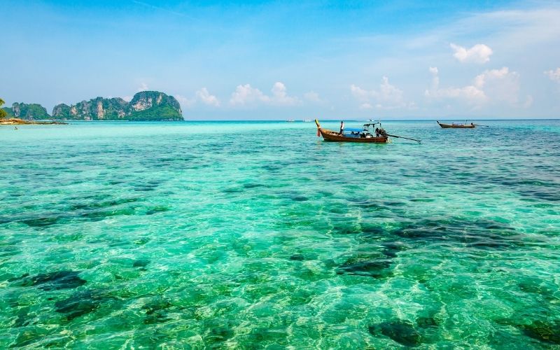 Clear turquoise waters and traditional long-tail boats floating near limestone cliffs on a sunny day at a tropical beach in Thailand.