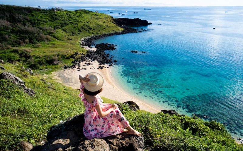 A panoramic view of Phu Quy Island showing crystal-clear turquoise waters, dramatic black volcanic rock formations, and colorful fishing boats floating peacefully near the shore under a bright blue sky.