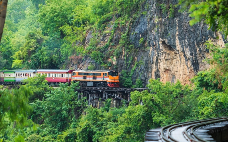 Wooden railway tracks of the Death Railway in Thailand, curving along limestone cliffs and dense green jungle landscape.