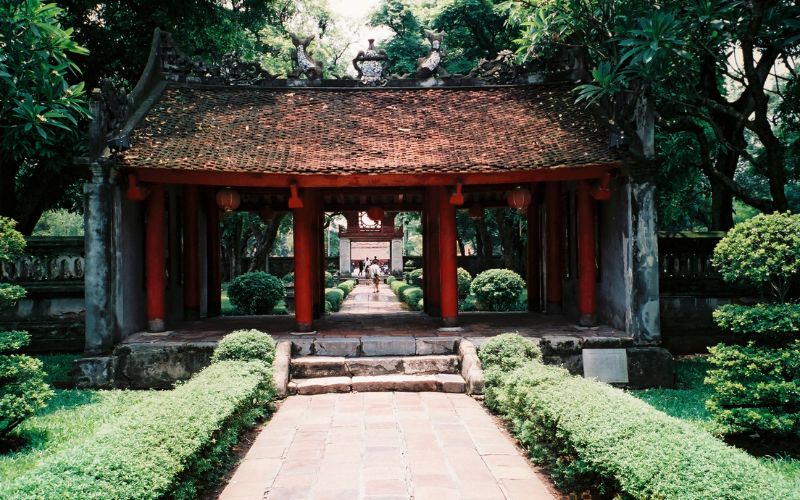A view of the Great Middle Gate (Dai Trung Mon) at the Temple of Literature. The traditional Vietnamese gate features a red-tiled roof with sweeping eaves and three entrances, flanked by two smaller side gates. It is surrounded by manicured green lawns and ancient trees under a soft, natural light.