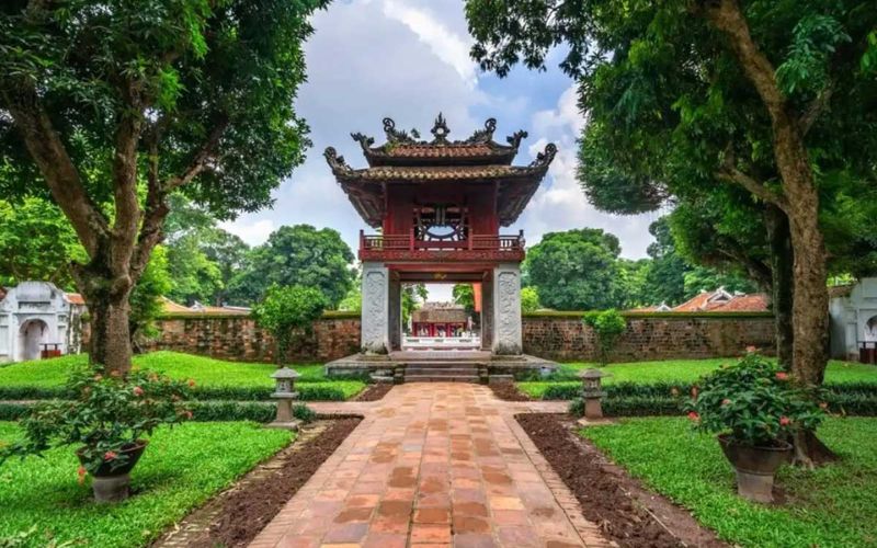 The iconic Khue Van Pavilion (Pavilion of the Constellation of Literature) at the Temple of Literature. The red wooden structure sits atop four white stone pillars and features a unique circular window representing a shining star. The pavilion is framed by greenery and ancient brick pathways.