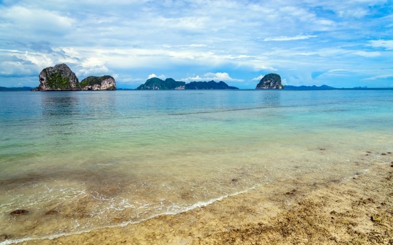 A wide scenic view of the ocean with waves gently washing onto a sandy shore in the foreground, and several large, rugged limestone karst islands dotting the horizon under a partly cloudy blue sky.