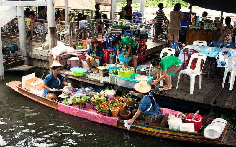 Taling Chan Floating Market in Bangkok with vendors selling food and produce from boats beside a riverside dining area