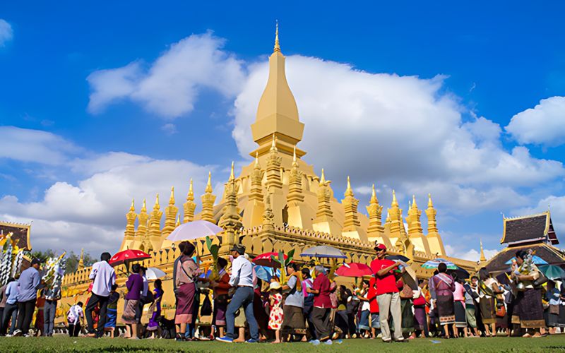 That Luang Festival in Vientiane, Laos, with devotees and monks participating in colorful religious ceremonies around the golden Pha That Luang stupa under a bright sky.