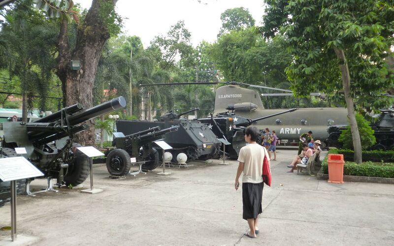 An outdoor display at the War Remnants Museum in Ho Chi Minh City, featuring several pieces of heavy military equipment including artillery guns, armored vehicles, and a large Chinook helicopter parked in a paved courtyard surrounded by trees.