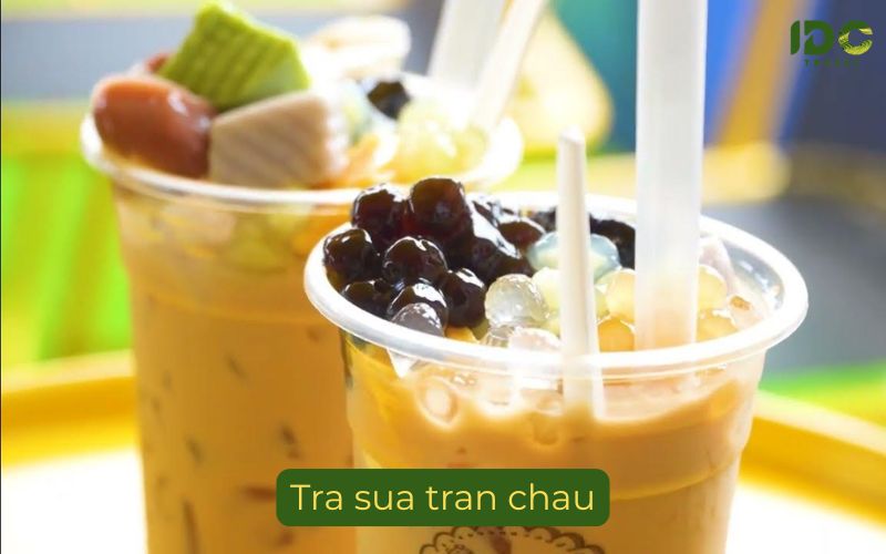 A close-up photograph of two plastic takeaway cups filled with bubble milk tea. The foreground cup is loaded with large, dark tapioca boba pearls and some smaller, clear bubbles, with a white straw inserted. The background cup shows toppings including a green jelly square and red fruits. The milk tea has a light brown, creamy appearance. A label reads "Tra sua tran chau" with the IDC Travel logo in the corner.