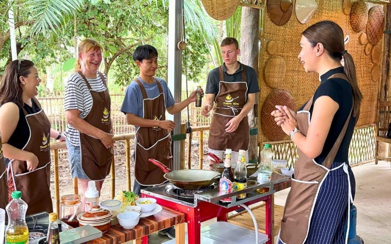 A group of five people, including a cooking instructor, participating in a Thai cooking class held in an open-air, rustic kitchen setting. Participants are wearing brown aprons and standing around a small cooking station with woks and ingredients. The instructor on the right is addressing the group, who are preparing to cook their dishes.