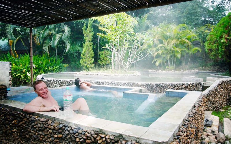 Tourists relaxing in natural hot spring pools at San Kamphaeng Hot Springs in Chiang Mai, Thailand, surrounded by lush tropical greenery and steam rising from the water