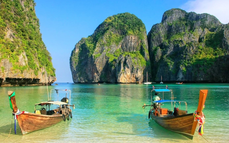 Colorful long-tail boats on the sandy beach of Koh Phi Phi, with limestone cliffs and clear blue water in the background.