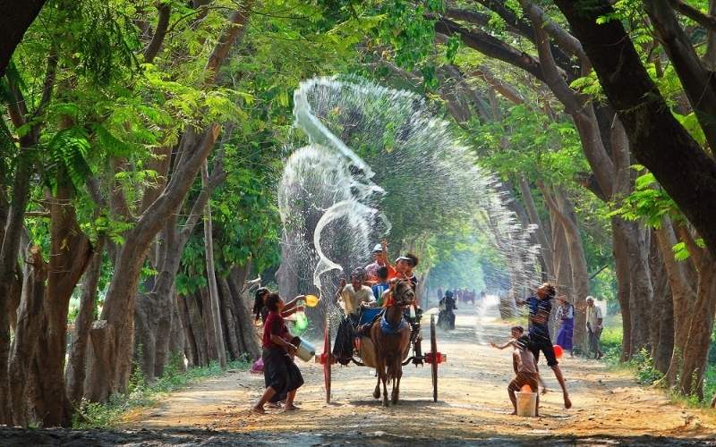 People celebrating the Thingyan Water Festival in Myanmar, splashing water on each other and on a passing horse cart under a canopy of green trees.