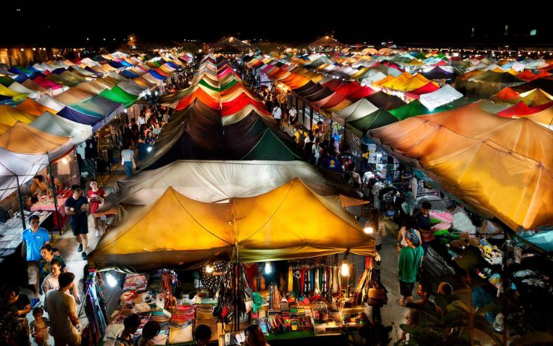 Aerial night view of Talad Rot Fai Market in Bangkok, Thailand, featuring colorful tents, vibrant lights, and bustling food and vintage stalls