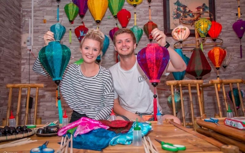 Two travelers smiling and holding colorful handmade lanterns during a traditional lantern-making workshop in Hoi An, Vietnam. The background is decorated with vibrant paper lanterns in various colors and shapes.