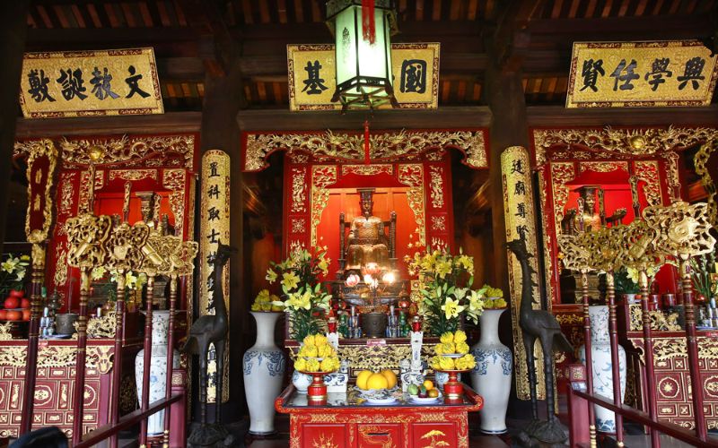 A view of the Fifth Courtyard at the Temple of Literature, featuring the Thai Hoc House. The large traditional wooden building has a dark-tiled, multi-tiered roof with ornate carvings along the ridges. It sits on a stone platform with wide steps, facing a spacious courtyard paved with bricks. The architecture reflects the classic Imperial style of the Le and Nguyen dynasties, surrounded by verdant trees.