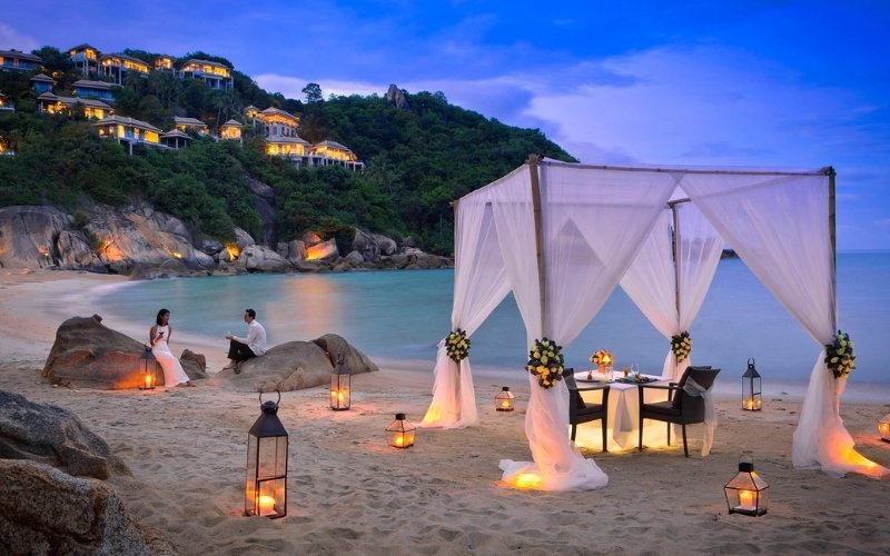 Romantic beachside dinner setup in Koh Samui, Thailand, with candlelit lanterns, a canopy draped in white fabric, and a couple sitting by the shore at sunset.