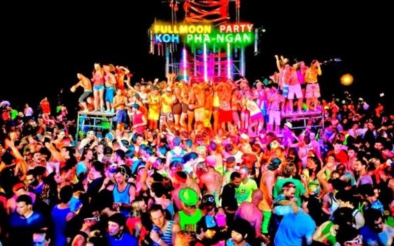 Crowds dancing on Haad Rin Beach during the Full Moon Party in Koh Phangan, Thailand, with neon lights and fire shows along the shoreline.