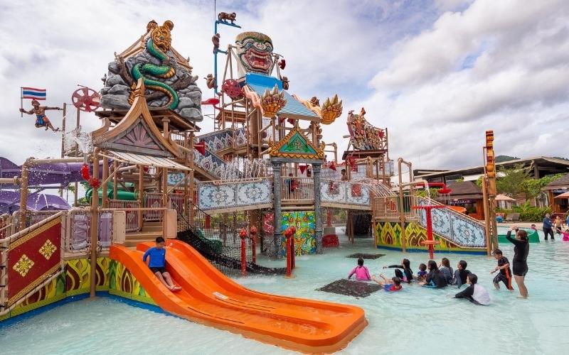 Children and families enjoy colorful water slides and play structures at a Thai-themed water park decorated with traditional statues and temple-style designs.