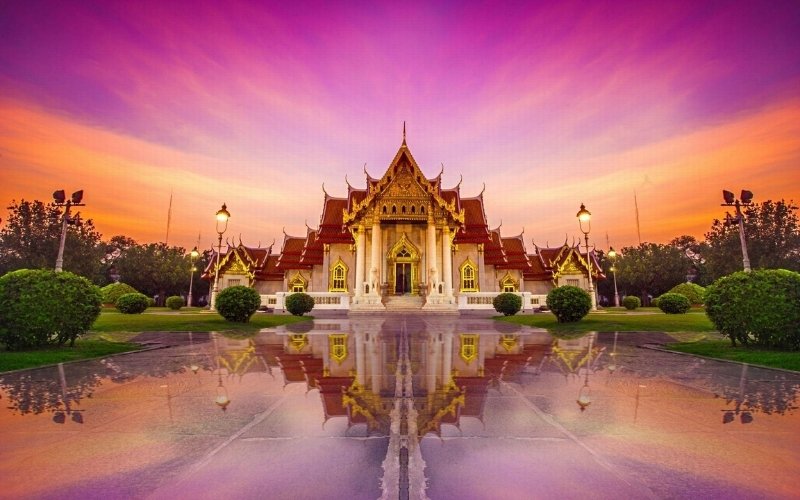 A beautifully illuminated Thai temple with ornate golden details, reflected on a wet marble courtyard under a vibrant purple and orange sunset sky.