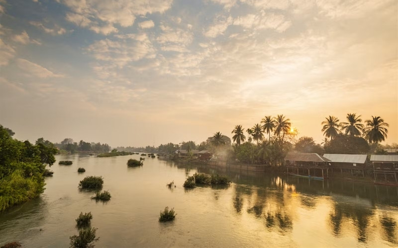 A tranquil riverside scene at sunset with golden reflections on the Mekong River, palm trees, and traditional riverside houses under a cloudy sky.