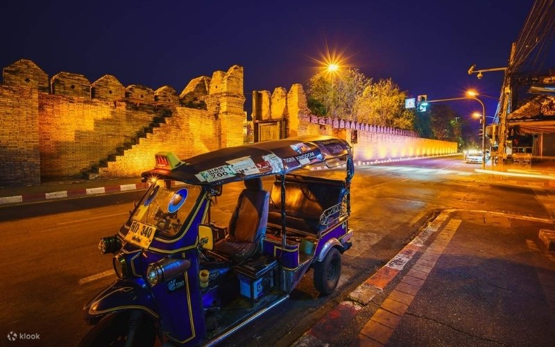 A colorful blue and purple tuk-tuk parked on a city street at night in Chiang Mai, Thailand, illuminated by warm streetlights. In the background, the ancient, yellow-orange brick wall of the Old City (Thapae Gate area) stretches along the street, creating a dramatic historical backdrop.