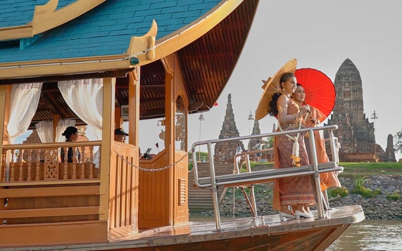Two women wearing traditional Thai costumes and holding parasols stand on the deck of a wooden boat cruising past the riverside ruins of Wat Chaiwatthanaram in Ayutthaya.