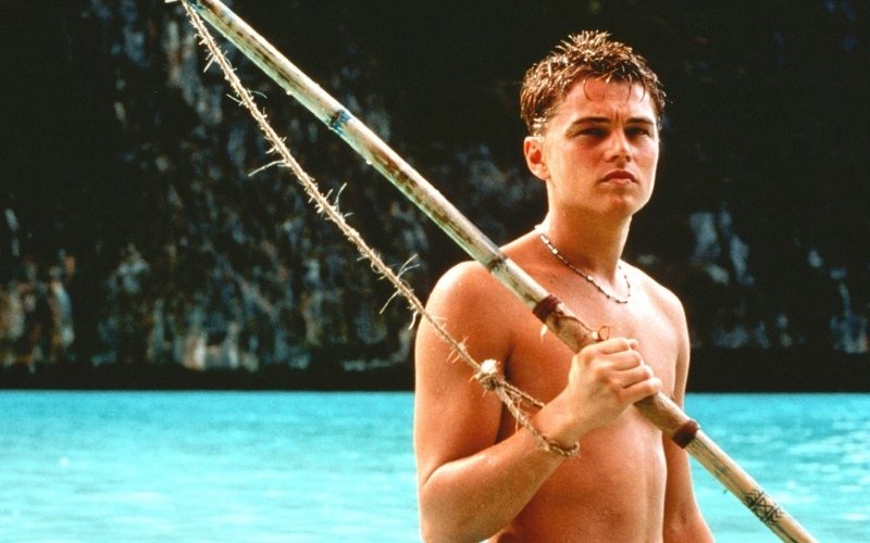 A young man standing in turquoise water holding a makeshift spear, with limestone cliffs and a tropical beach in the background, from a scene filmed at Phi Phi Island, Thailand.
