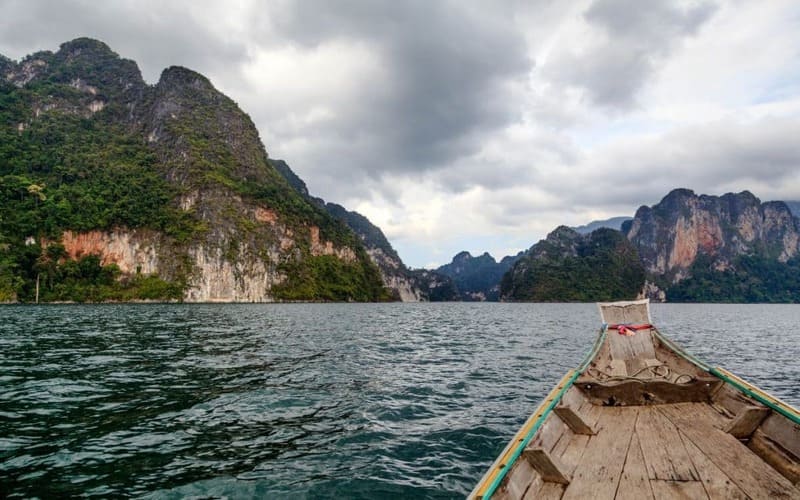 Wooden long-tail boat cruising across Cheow Lan Lake in Khao Sok National Park, surrounded by dramatic limestone cliffs and lush rainforest.