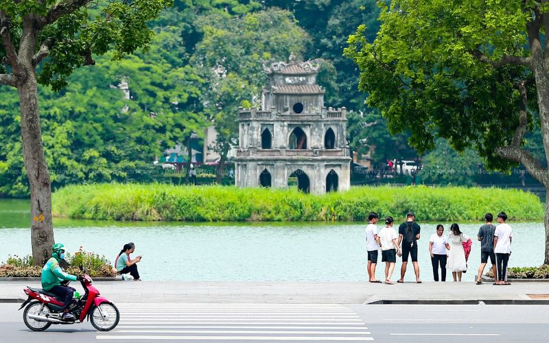 A daytime view of the Hoan Kiem Lake promenade in Hanoi, featuring people walking and standing near the water's edge with the historic Turtle Tower visible on a small green island in the background.