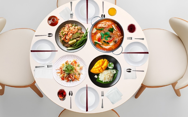 A round dining table set for four with traditional Thai dishes including pad thai, tom yum soup, som tam (papaya salad), and mango sticky rice, surrounded by neatly arranged plates, glasses, and utensils.