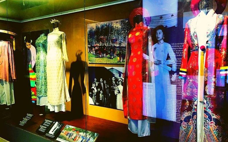 A well-lit gallery space showcasing a diverse collection of traditional Vietnamese Ao Dai. Mannequins are dressed in various styles of the national garment, ranging from classic, simple designs to more ornate, colorful versions with intricate embroidery, displayed behind protective glass and alongside informational plaques.