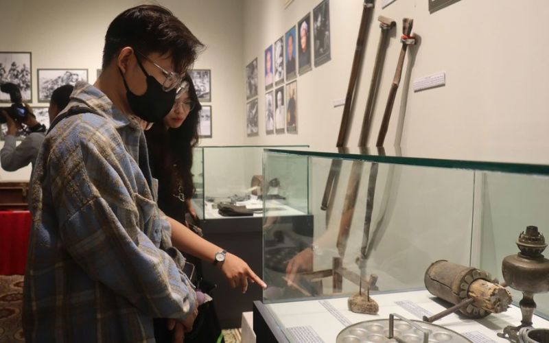 A display of historical artifacts from the war era at the Southern Women's Museum. The collection includes everyday items used by female soldiers and resistance fighters, such as canteens, sandals made from tires, woven scarves (Krama), and various hand-held tools, all presented in a glass display case with historic black-and-white photos in the background.