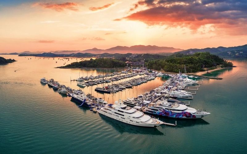 An aerial, wide-angle photo of a picturesque, horseshoe-shaped marina at sunset, filled with numerous large white luxury yachts and smaller vessels, set against a backdrop of green tropical hills and calm bay water.