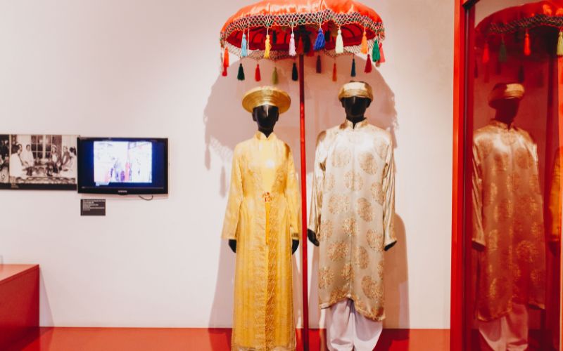An interior view of a modern gallery hall within the museum used for rotating exhibitions. The space features large-scale photographic prints and artistic installations suspended from the ceiling, with visitors walking through the open-plan area to view displays focused on contemporary women's issues and traditional crafts.