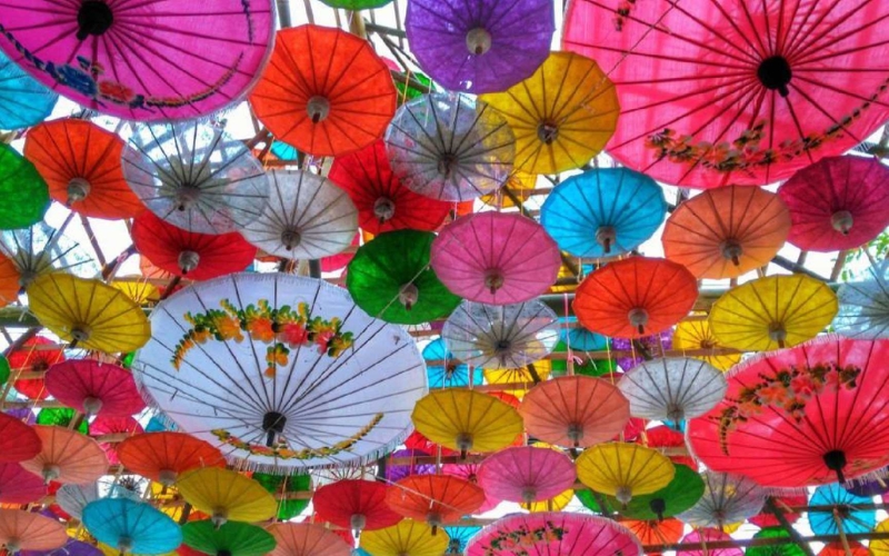 A nighttime photo of a brightly lit bridge over a river, decorated with neon lotus flower designs and linear blue and pink lights, while a crowd of people watches from the side, with some holding phones to take pictures.