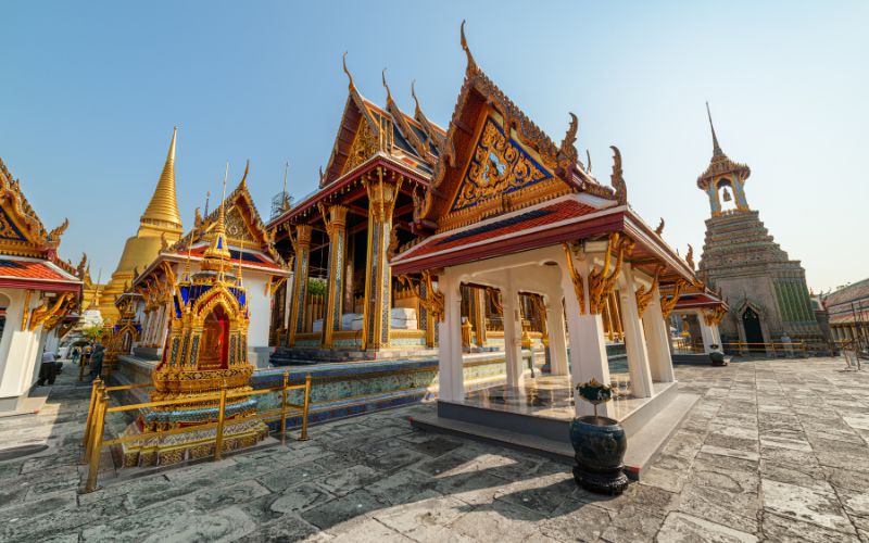 A ground-level view from an uneven stone courtyard, looking across the main terrace toward the gilded structure of the ubosot, the golden chedi, and the tiered bell tower (Hor Rakang) under a bright, sunny sky.