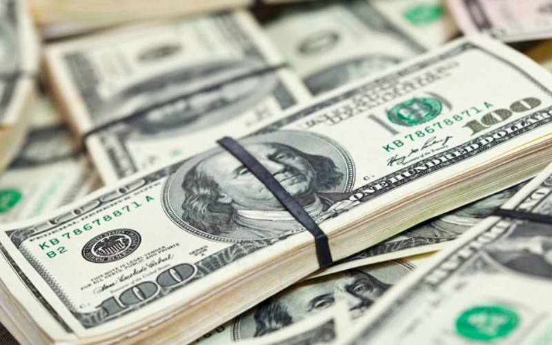 A stack of 100 USD bills bound with a black rubber band, placed on a background of other USD notes.