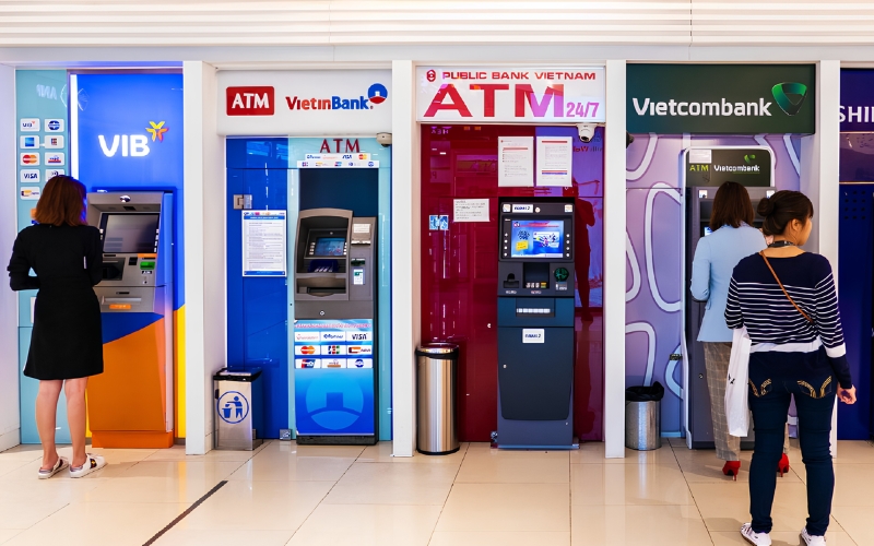 Row of ATM machines from different banks including VIB, VietinBank, Public Bank Vietnam, and Vietcombank, with people withdrawing cash.
