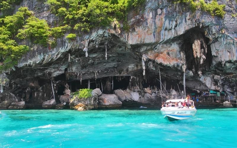 A speedboat carrying tourists approaches Viking Cave on Phi Phi Island, Thailand, surrounded by turquoise water and steep limestone cliffs covered in greenery.