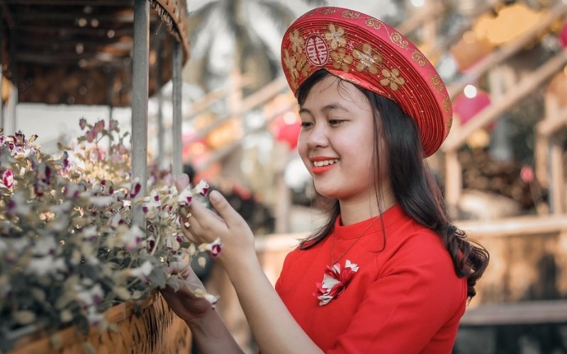 A young woman wearing a traditional Vietnamese red áo dài and decorative headdress smiling while arranging flowers at a cultural festival.