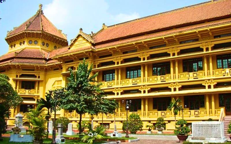 A side-angle view of the Vietnam National Museum of History in Hanoi, showcasing its distinctive Indochina architectural style with yellow walls, red-tiled tiered roofs, and an octagonal pavilion under a clear sky.